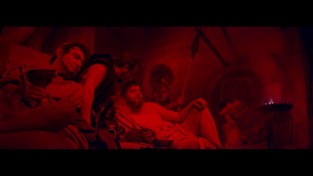A scene depicting several men in ancient attire sitting in a dimly lit, red-hued environment, with one man reclining and others engaged in conversation, highlighting a historical or mythological theme.