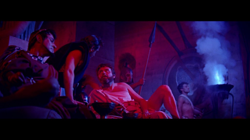 A dramatic scene depicting ancient Roman or Greek characters with vibrant red and blue lighting. Several men are engaged in conversation, dressed in togas and armor, while one is seated and holding a bowl. A figure in the background operates a fire source, contributing to the atmospheric lighting.