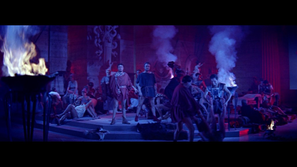 A dramatic scene set in an ancient Roman environment, with individuals dressed in historical garments, surrounded by torches and smoke, depicting a gathering or ceremony.