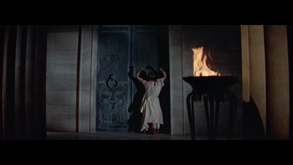 A figure in a draped garment is seen pushing against a large, ornate door, with a flickering flame from a nearby torch illuminating the scene.