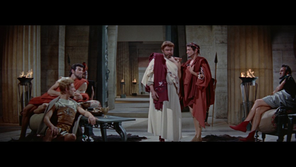 A group of ancient Roman characters in a dramatic scene set in a grand hall, featuring men dressed in togas and robes, with some seated and others engaging in conversation.