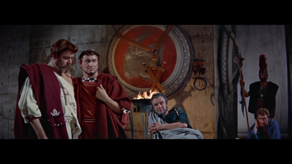 A scene depicting four men in ancient Roman attire indoors, with a large decorative wall behind them. One man is seated, while others stand and appear to be engaged in a serious discussion.