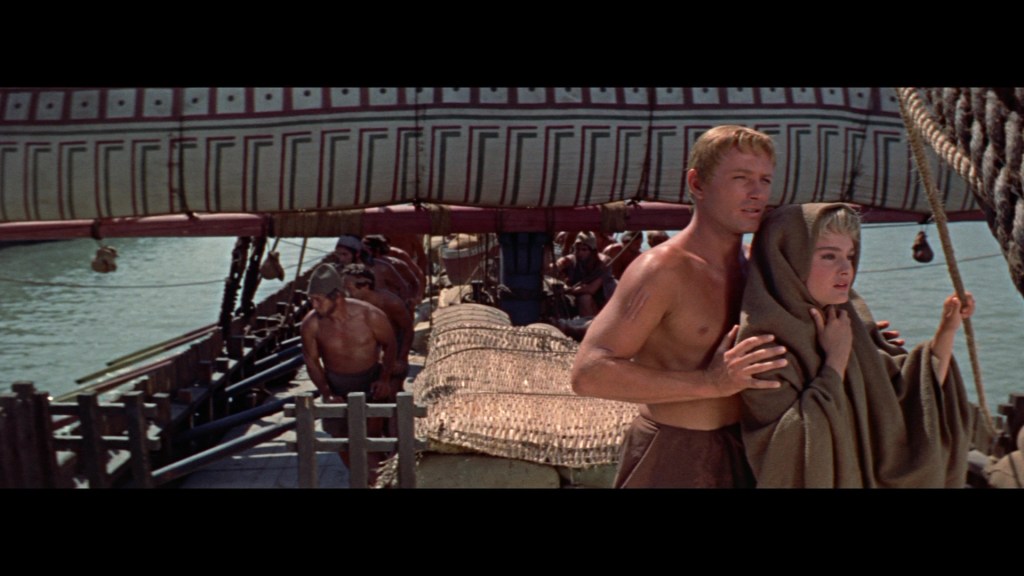 A scene depicting two characters on a ship, with one man holding a woman closely. The ship features an ornate sail and other crew members visible in the background, suggesting a historical or epic setting.
