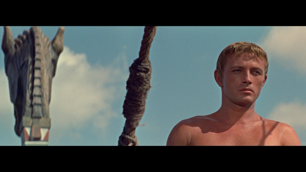 A shirtless man stands in front of a towering horse statue against a blue sky.