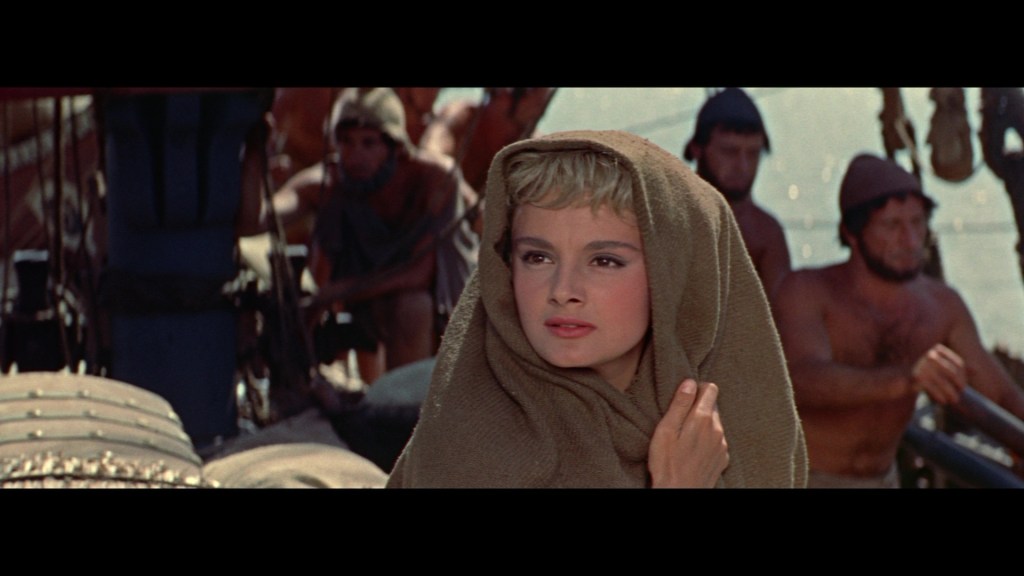 A young woman draped in a shawl stands on a ship, gazing thoughtfully, with sailors working in the background.