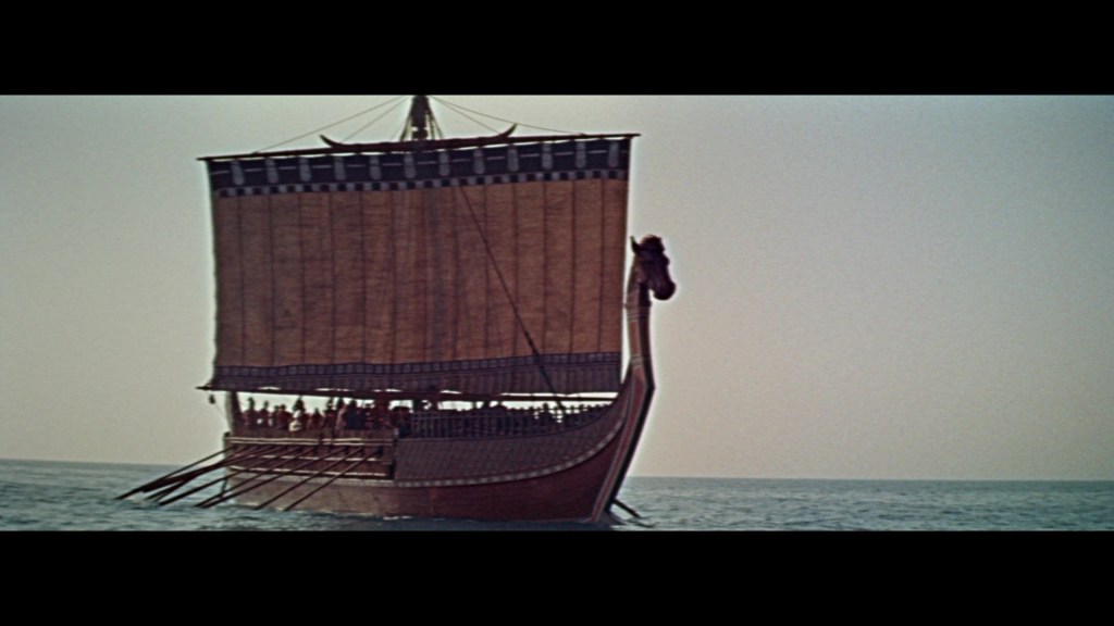 A large ancient ship with a prominent figurehead, sailing on calm waters under a clear sky.