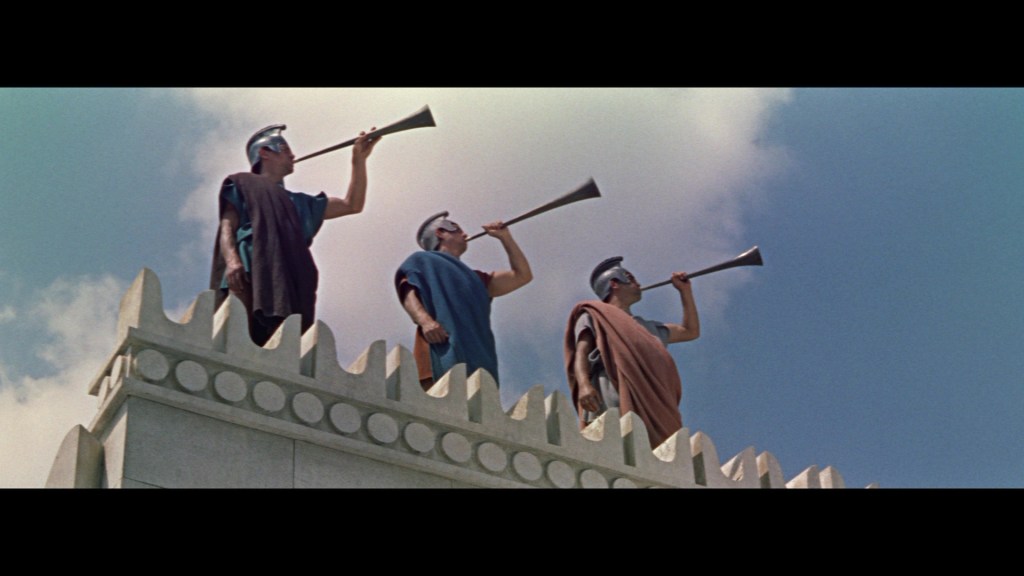 Three ancient Roman soldiers in tunics and helmets blowing horns from a tall structure under a blue sky.