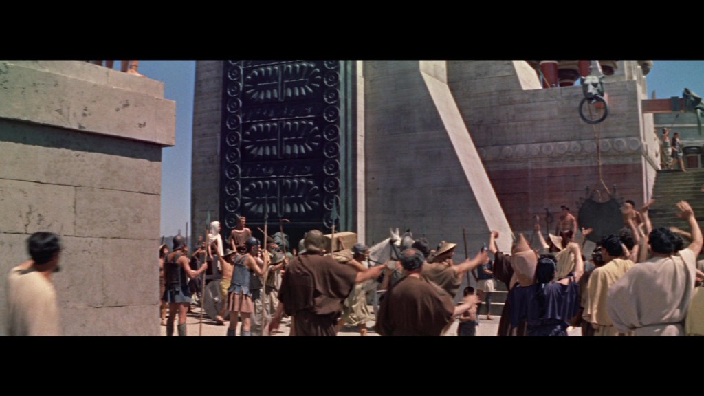 A large crowd of ancient Greek citizens gathered in front of a monumental structure, with some people raising their arms in excitement. The scene depicts a lively atmosphere, suggesting a public event or celebration.