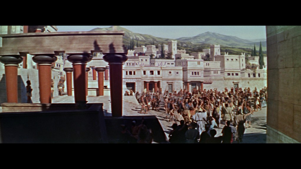 A bustling scene depicting an ancient city with people gathered in a square, featuring traditional architecture and mountainous scenery in the background.
