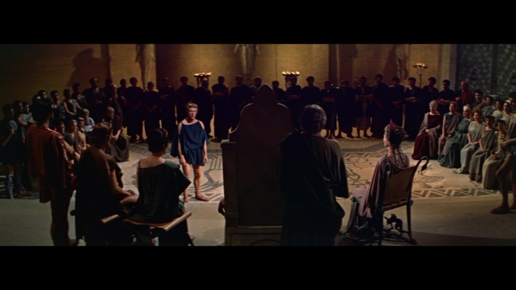 A dramatic scene depicting a young man standing in front of an audience in ancient attire, with figures seated in a semicircle, in a grand hall illuminated by candlelight.