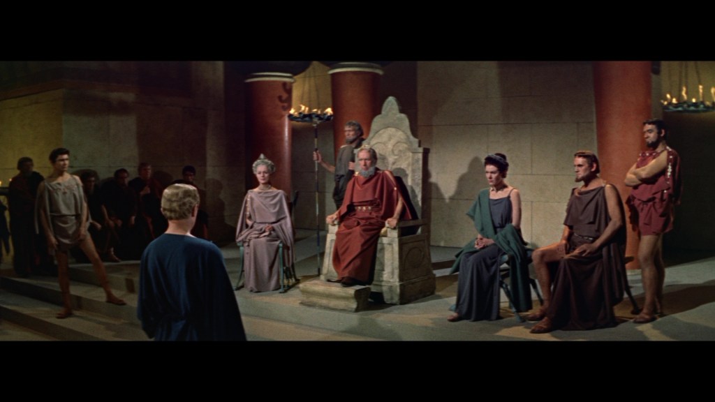 A dramatic scene depicting a group of figures dressed in ancient Roman attire, seated in a grand setting. In the foreground, a man in a blue robe stands before a throne occupied by a dignified figure. Surrounding them are additional characters sitting on either side, dressed in various colors of robes, with an audience in the background.