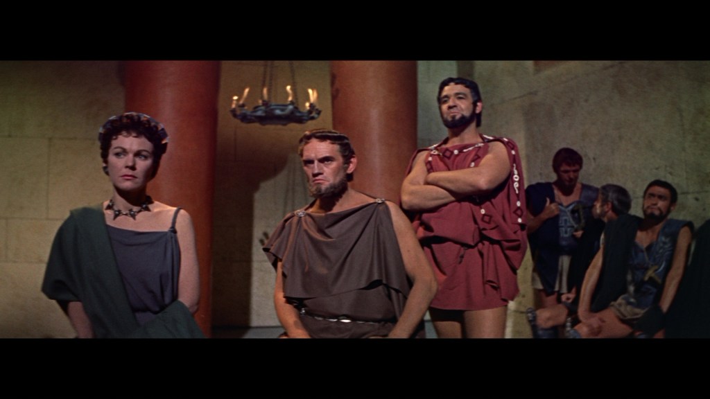 A group of ancient Roman or Greek characters, dressed in traditional togas, are standing in a stone chamber. The scene features a woman with dark hair wearing a headdress, a man with a beard in a brown toga, and several other men in various outfits conversing in the background.