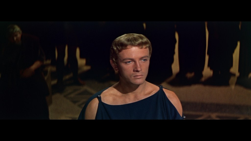 A young man in a blue toga stands facing the camera, with a serious expression, surrounded by blurry figures in dark clothing.