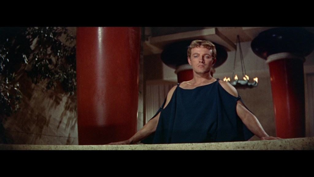 A man wearing a dark toga stands on a balcony, overlooking a room with red columns and a chandelier in the background.