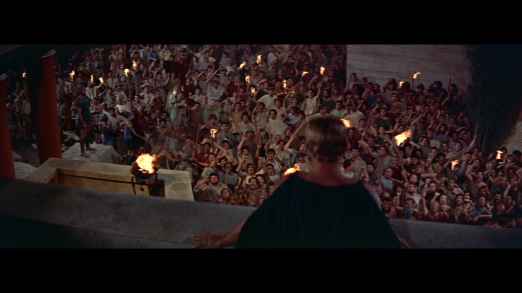 A large crowd of people holding torches gathers in front of a speaker on a raised platform, creating a dramatic atmosphere.