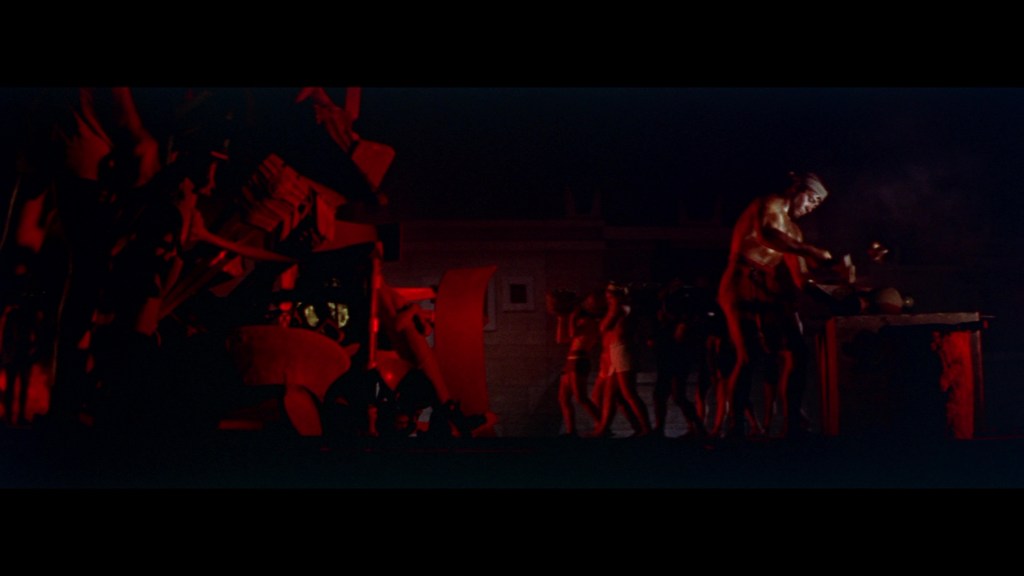 A dimly lit scene with red lighting, featuring several figures engaged in a ritualistic activity, some holding pots and other objects, while one figure is working at a table.