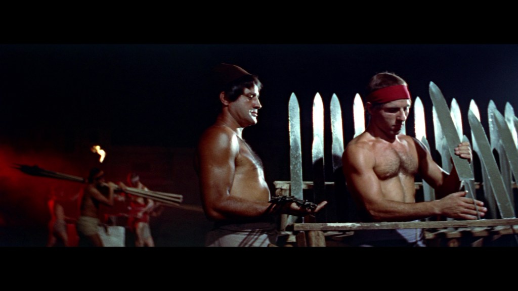 Two muscular men are seen working together to assemble weapon-like structures in a dimly lit setting, with other figures in the background preparing for an activity.