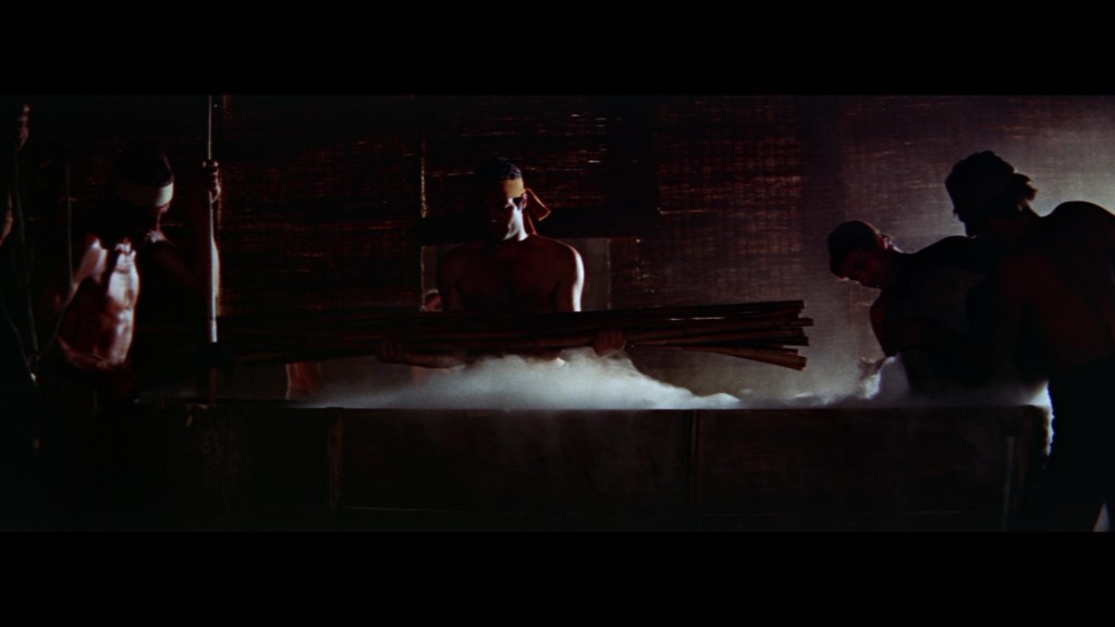 A group of shirtless men working in a dimly lit environment, surrounded by steam. They are holding wooden sticks and are engaged in a task near a large steaming vat.