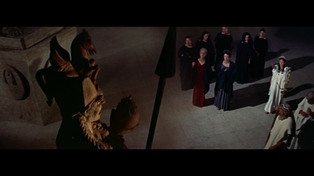 A group of women in dark and light robes gathered in a dramatic setting, looking up towards a large, ornate statue in a dimly lit space.