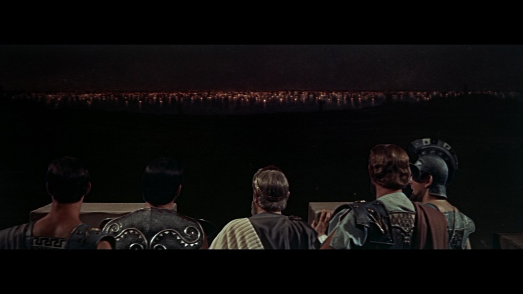 A group of ancient warriors and leaders stands with their backs to the viewer, gazing at a distant city skyline illuminated by lights at night.