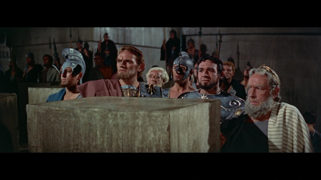 A group of ancient Roman men in togas and armor gathered in a large hall, observing a momentous event with serious expressions.