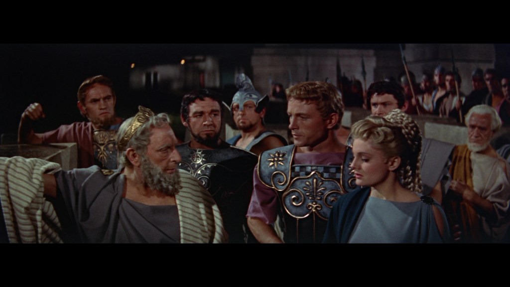 A group of ancient Greek characters in a dramatic scene, featuring a bearded man in regal attire, armored figures, and a woman in traditional dress, set against a backdrop of a crowd with spears.