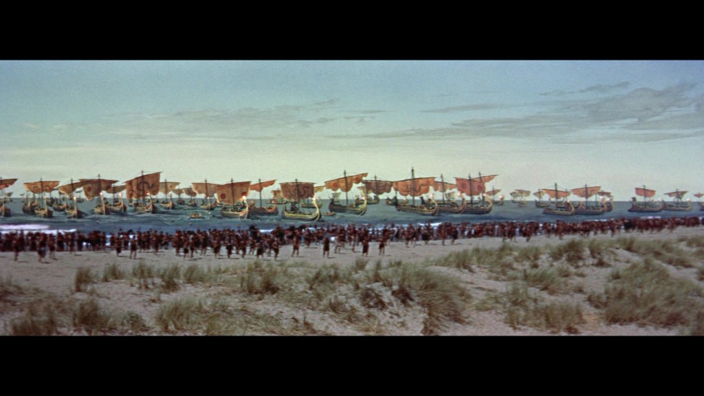 A large gathering of people on a sandy beach, watching a fleet of ancient ships with red sails approaching the shore.