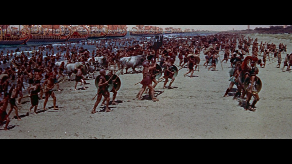A large group of ancient warriors in armor and shields gathered on a sandy beach, preparing for battle, with ships visible in the background.