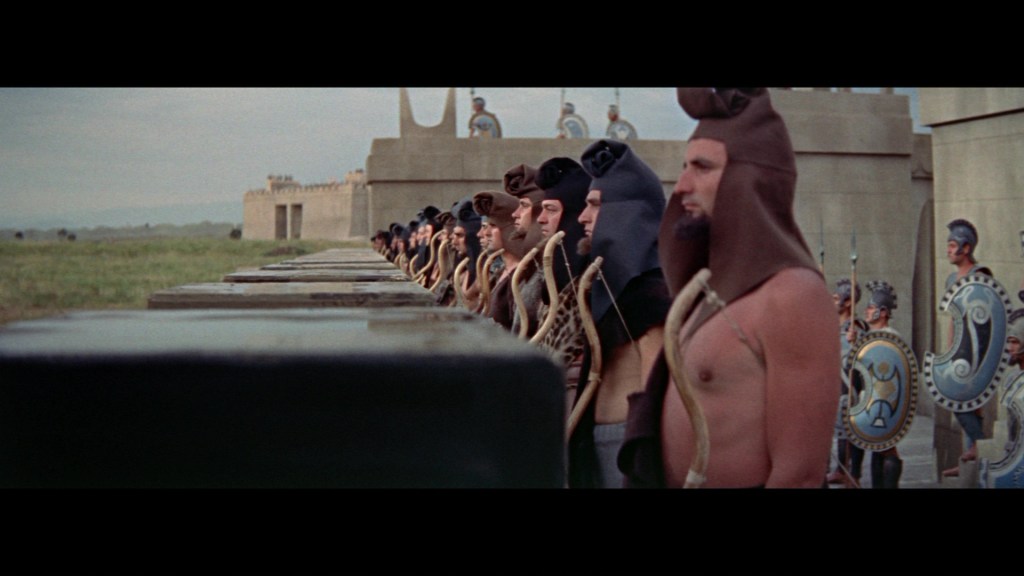 A line of ancient warriors wearing elaborately designed helmets and costumes, standing at attention in a dramatic landscape, with stone structures and soldiers in armor visible in the background.