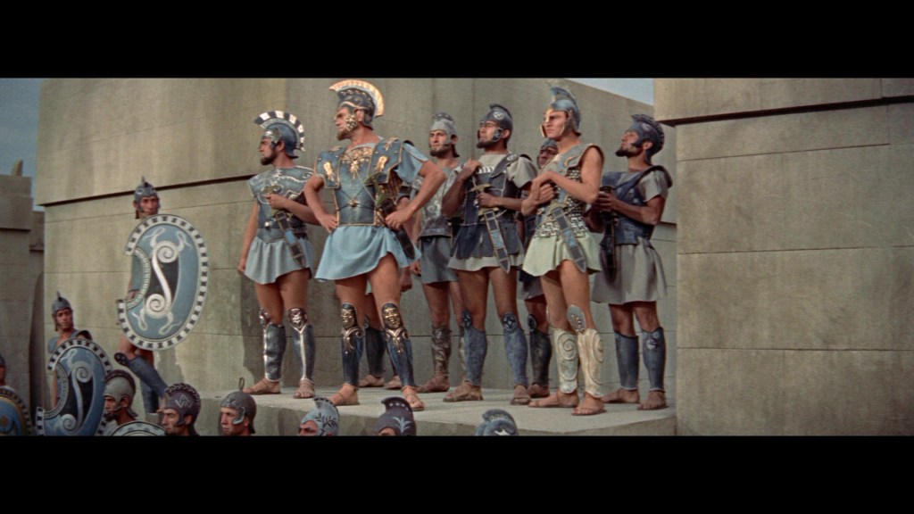 A group of ancient warriors dressed in Greek-style armor and helmets, standing on a stone platform, with decorative shields at their feet.