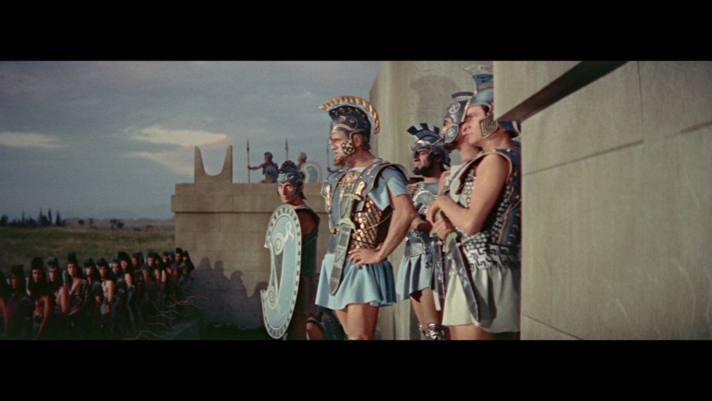 A line of ancient Greek soldiers in armor, standing on a fortress wall, observing a crowd below. A commanding figure in the center wears a decorative helmet and shield, while soldiers in tunics with armor stand beside him, set against a cloudy sky.