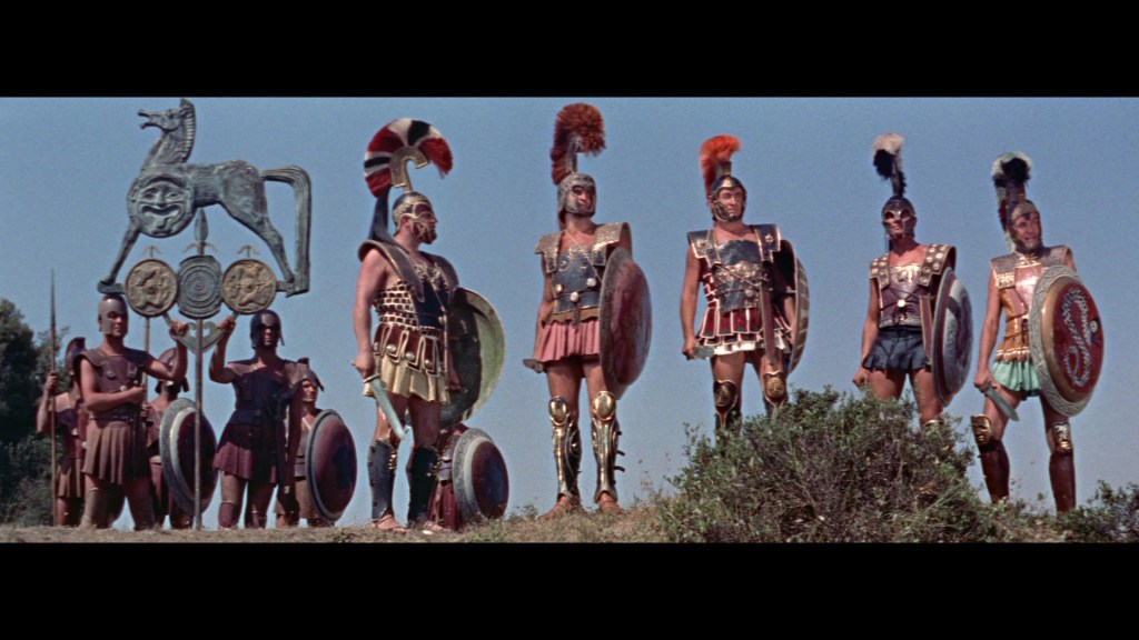 A group of ancient warriors in armor and helmets, standing in formation with shields and spears, in front of decorative banners on a hillside.
