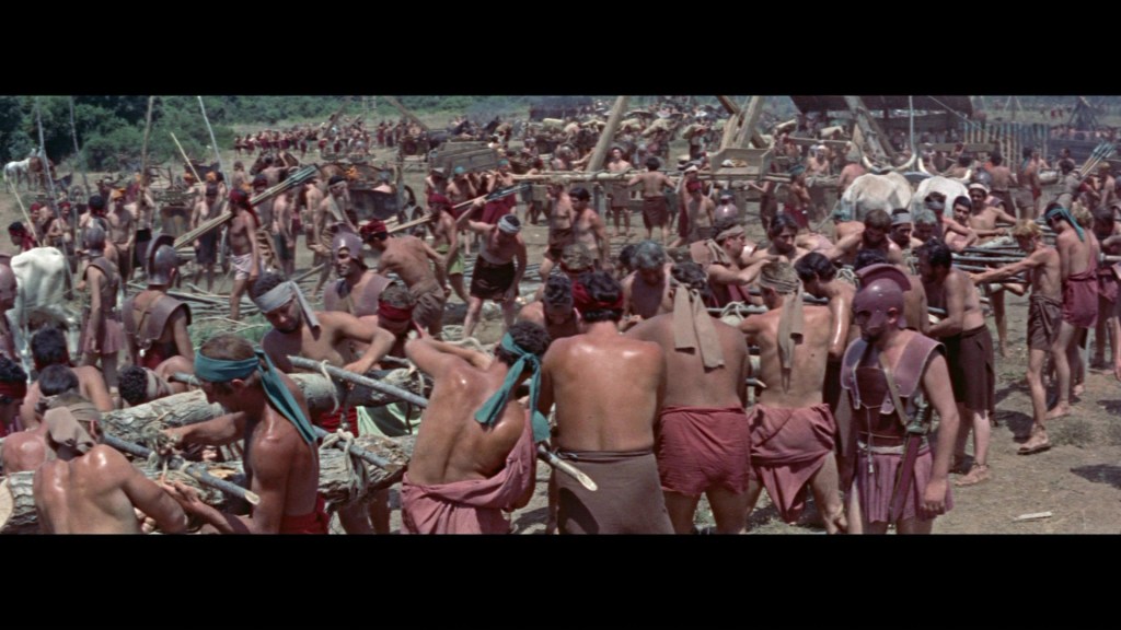 A large crowd of people in ancient attire are engaged in manual labor, constructing structures with wooden beams. Many men are bare-chested and working together in an outdoor setting, surrounded by greenery.