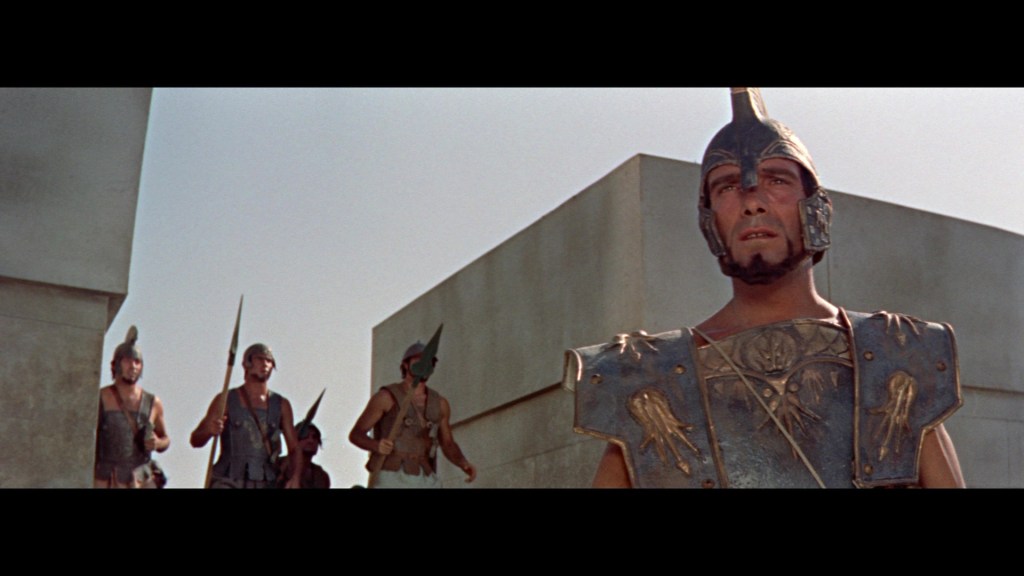 A group of armored warriors in ancient Greek attire standing near a stone structure, with one warrior prominently featured in the foreground wearing a helmet and intricate armor.