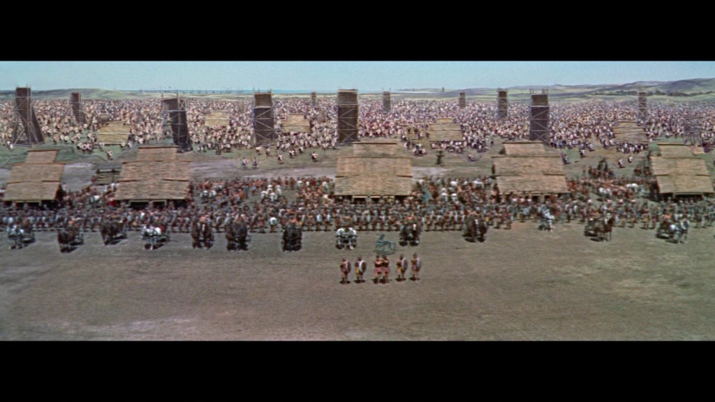 A wide aerial view of a large gathering of people in a vast open area, with numerous wooden structures and towers in the background.