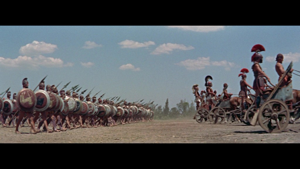 A scene depicting an ancient battle with rows of armored warriors holding shields and spears, lined up against chariots in a dry landscape under a blue sky.