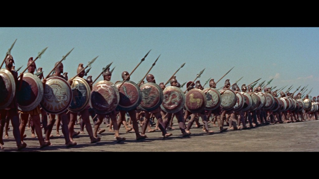 A formation of ancient soldiers marching in unison, each carrying a round shield and spear under a clear blue sky.