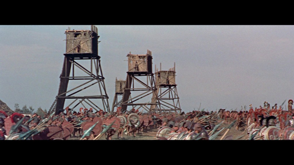A battlefield scene featuring wooden towers and soldiers in formation, with shields and spears, set against a clear sky.