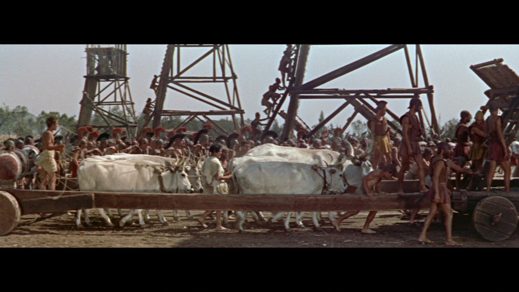 A large group of people, including workers and soldiers, are engaged in a construction scene with oxen pulling a wooden cart, set against a backdrop of structures resembling ancient machinery.