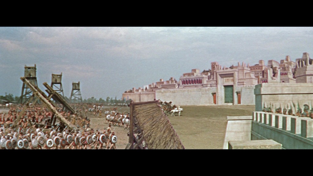 Ancient battlefield scene with soldiers and catapults, set against a large fortified city in the background.