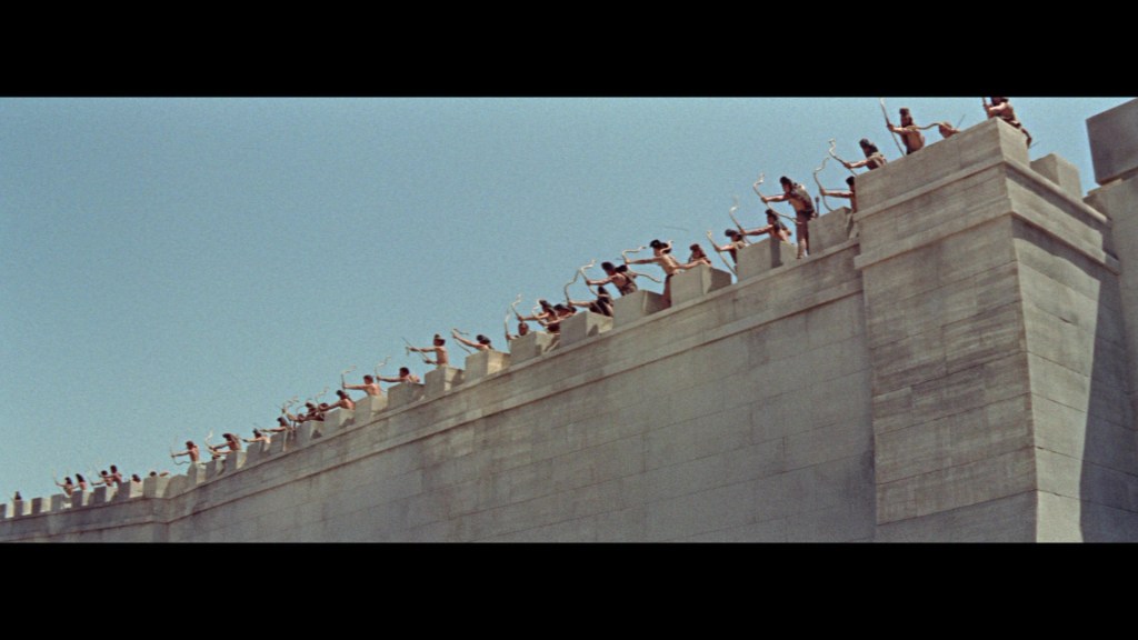 Line of archers standing on a fortress wall under a clear blue sky.