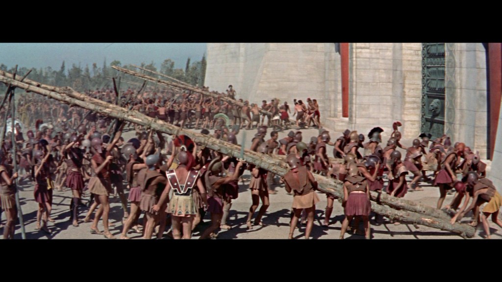 A large group of ancient warriors in short tunics and helmets are seen lifting and carrying long wooden logs, set against a backdrop of large stone walls with intricate carvings.