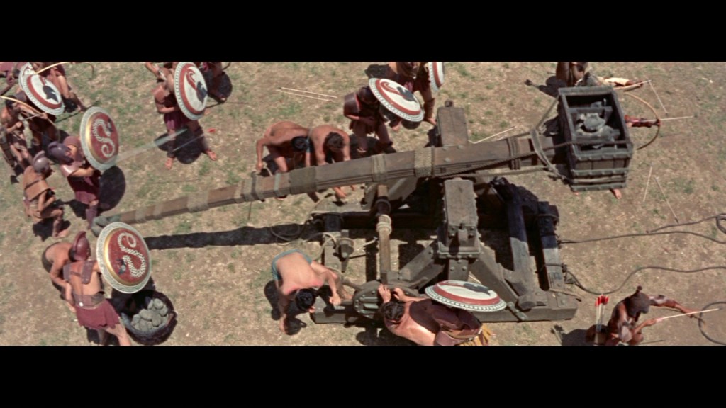 An overhead view of ancient warriors assembling a large catapult on grass, with some wearing shields and traditional attire.