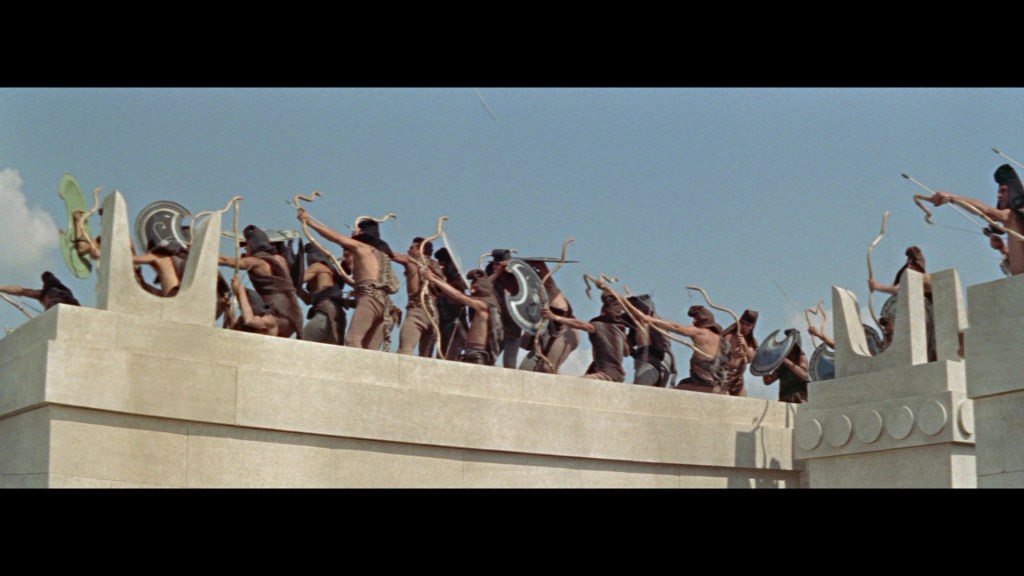 A scene depicting ancient warriors, armed with shields and bows, preparing to defend a high stone wall under a clear blue sky.