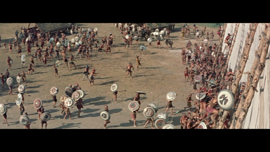 A large group of people in ancient attire engage in a battle scene, armed with shields and weapons, set in an open field surrounded by structures.