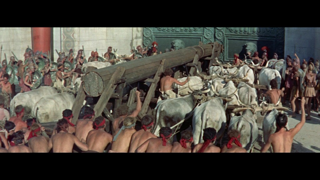 A large group of ancient soldiers and civilians gather around a wooden siege engine, drawn by oxen, in front of a large gate. Many individuals are wearing historical costumes, with some raising their arms in excitement.