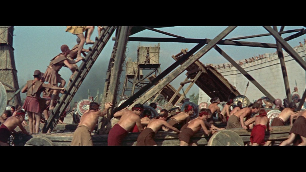 A scene depicting a large group of historical figures, possibly warriors, engaged in a siege. Some are climbing a ladder while others prepare to attack, showcasing a chaotic battle environment.