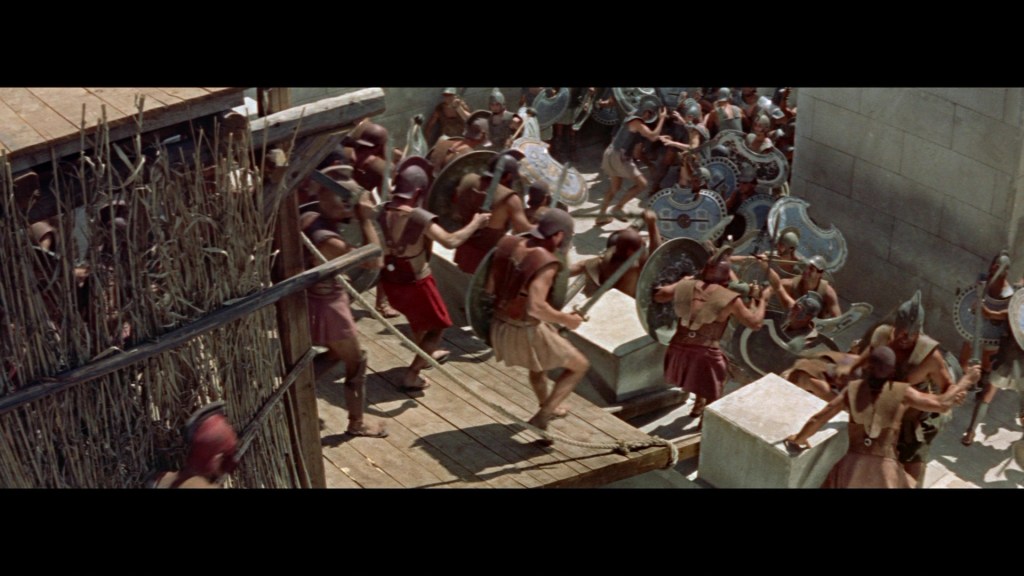 A large group of ancient soldiers engaged in battle, wearing armor and holding shields, with a siege tower in the foreground and a stone city wall in the background.