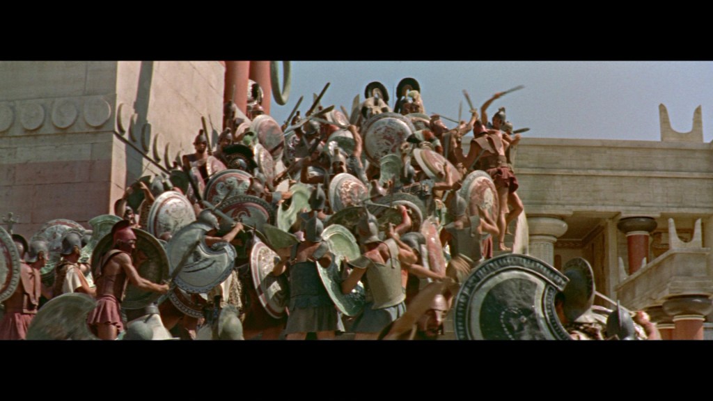 A group of armored warriors with shields, depicted in a historical battle scene, advancing towards a brick structure with columns.