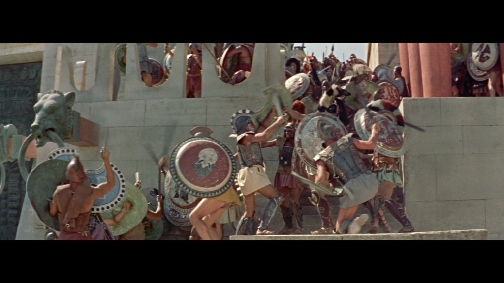 A scene depicting ancient warriors in battle, wearing armor and holding shields, some climbing stairs, with a large building structure in the background.